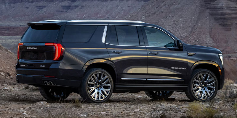 Rear-three-quarter view of a dark gray GMC Yukon Denali parked on rocky, dirt terrain with desert mountains in the background.