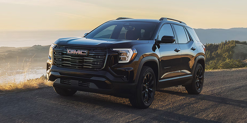 Front-three-quarter view of a black GMC Terrain driving along a scenic coastal ridge road at sunset.
