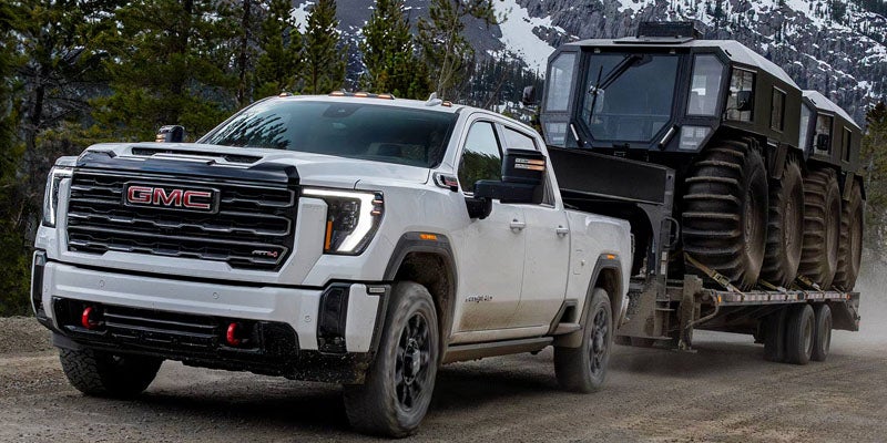 A white GMC Sierra HD AT4X pickup truck towing a large, multi-wheeled off-road vehicle on a flatbed trailer through a mountain landscape.
